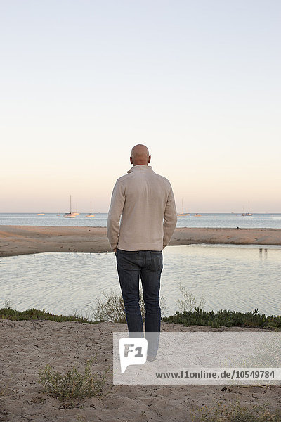 Bald man standing on a sandy beach by the ocean.