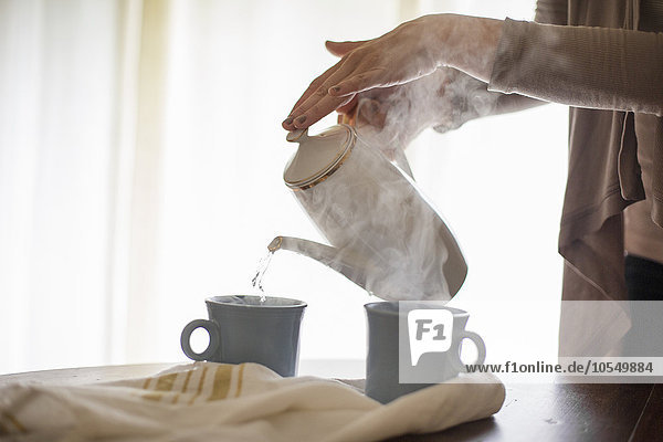 Nahaufnahme einer Frau  die heißes Wasser aus einer Kaffeekanne in eine Tasse gießt.