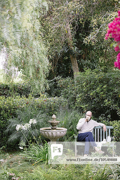 Mann sitzt auf einer Holzbank in einem Garten und macht eine Pause.
