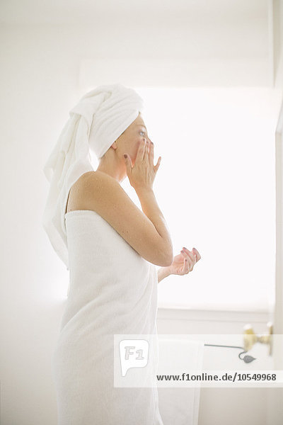 In ein weißes Handtuch gewickelte Frau  die in einem Badezimmer steht und ihr Gesicht eincremt.