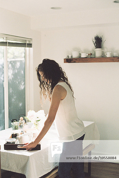 Eine Frau stellt ein Tablett mit einer Teekanne und einer Vase mit weißen Rosen auf einen Tisch.