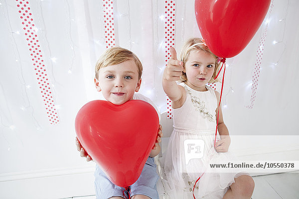 Young boy and girl posing for a picture in a photographers studio  holding red balloons.