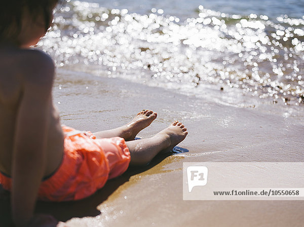 Ein Junge sitzt auf dem Sand am Wasser und beobachtet  wie die Wellen den Strand erreichen.