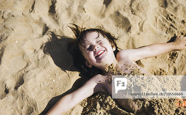 A boy lying on his back on the beach  his torso covered in sand  laughing.