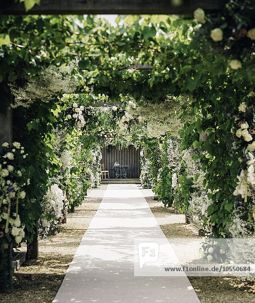 A garden path through an arch leading to an alcove with table and chairs with climbing plants and white roses.