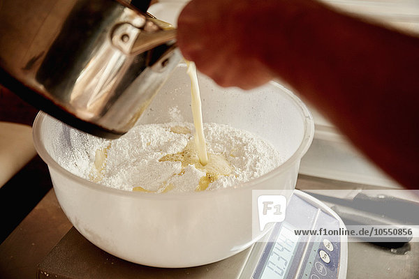 Ein Bäcker gießt Flüssigkeit aus einem Kupferkrug in eine Schüssel mit Mehl und Zutaten für den Teig.