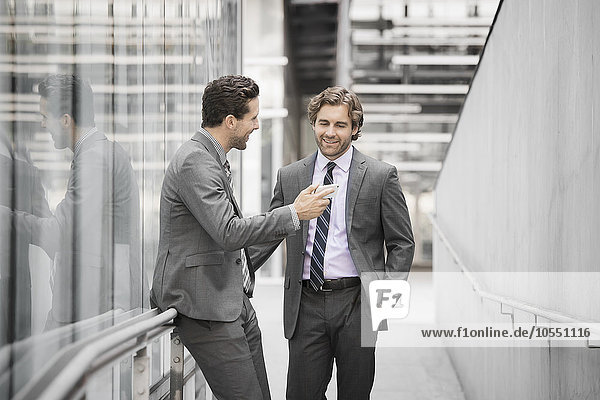 Zwei Männer in Geschäftsanzügen vor einem großen Gebäude  einer mit einem Smartphone in der Hand.