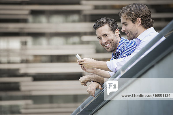 Zwei Männer lehnen an einem Geländer  einer hält ein Smartphone.