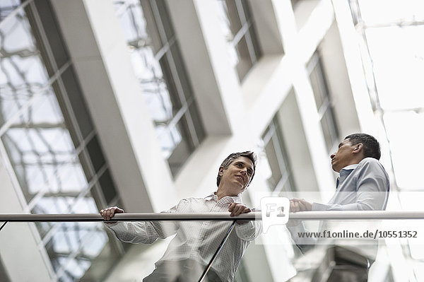 Zwei Männer  Geschäftskollegen  stehen an einem Geländer in einem Atrium oder Innenhof und unterhalten sich.