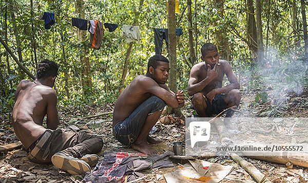 Drei junge Männer der Ureinwohner Orang Asil sitzen auf dem Boden im Dschungel und rauchen  indigenes Volk  tropischer Regenwald  Nationalpark Taman Negara  Malaysia  Asien