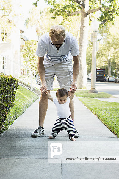 Father helping baby son walk on sidewalk