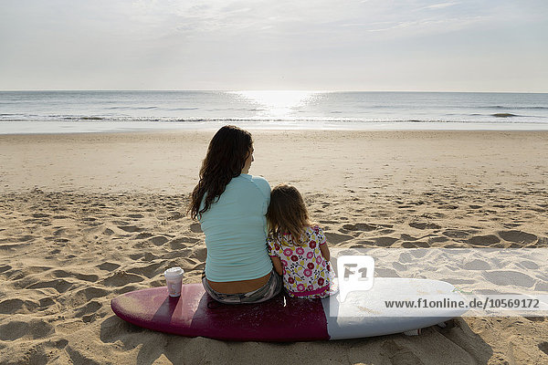 Mutter und Tochter sitzen auf einem Surfbrett am Strand