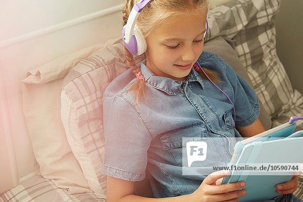 Ausschnitt eines Mädchens  das mit Kopfhörern auf dem Bett sitzt und lächelnd auf ein digitales Tablett blickt.