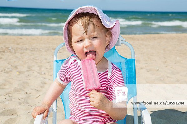 Weibliches Kleinkind trägt Sonnenhut und isst Eis am Strandkorb.