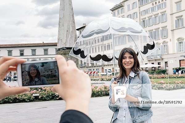 Young woman holding umbrella posing for friend using smartphone to take photograph  Piazza Santa Maria Novella  Florence  Tuscany  Italy