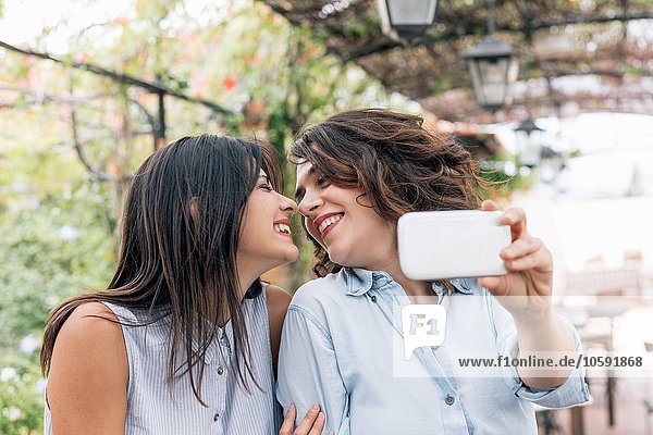 Lesbian couple in plant covered archway using smartphone to take selfie face to face  Florence  Tuscany  Italy