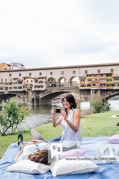 Lesbian couple on blanket using smartphone to take photograph in front of Ponte Vecchio and river Arno  Florence  Tuscany  Italy