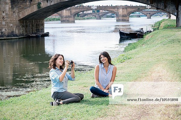 Lesbian couple sitting on arno river bank in front of Ponte Vecchio holding digital camera smiling  Florence  Tuscany  Italy