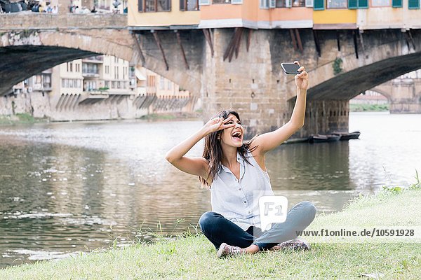 Young woman sitting on riverbank in front of Ponte Vecchio and Arno river using smartphone to take selfie  Florence  Tuscany  Italy
