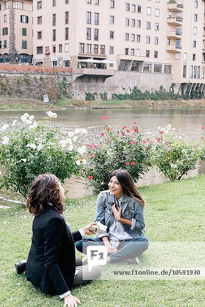 Lesbian couple sitting on grass next to Arno river giving  receiving gift  Florence  Tuscany  Italy
