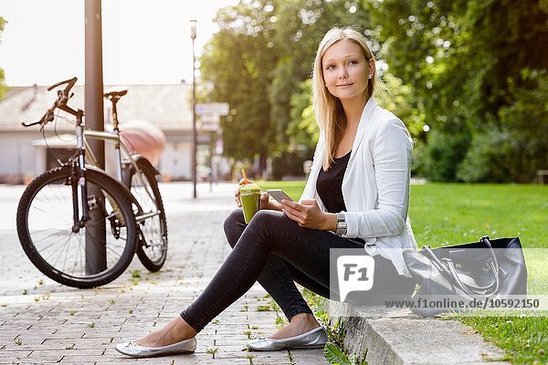 Junge Frau mit Smoothie-Drink und Smartphone im Park sitzend