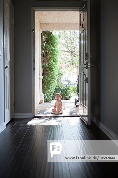 View through entrance hall of baby boy sitting outside at open front door  looking at camera open mouthed