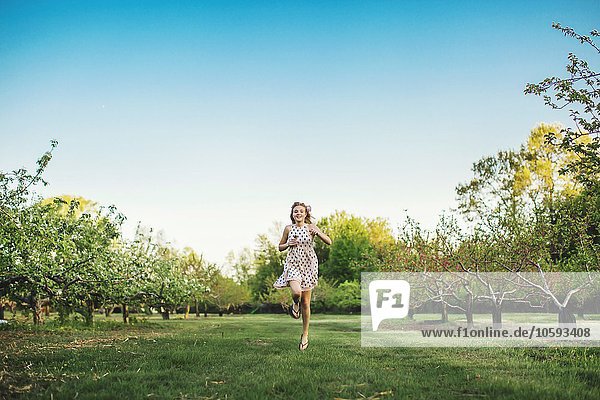 Full length front view of young woman wearing sleeveless dress running through orchard  looking at camera smiling