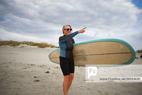 Seniorin steht am Strand  hält Surfbrett  zeigt auf das Meer