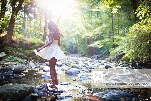 Glamouröse junge Frau in weißem Kleid  die über die Felsen des Waldes tritt.