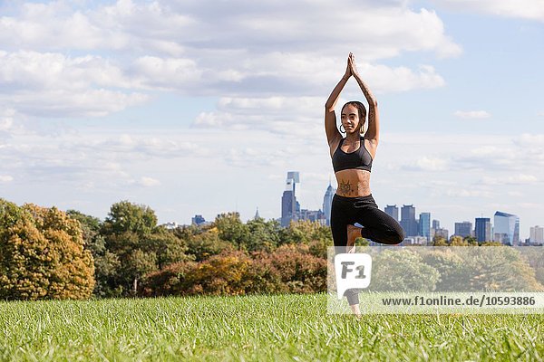 Full length front view of young woman on grass in yoga position  on one leg  arms raised  hands together  Philadelphia  Pennsylvania  USA