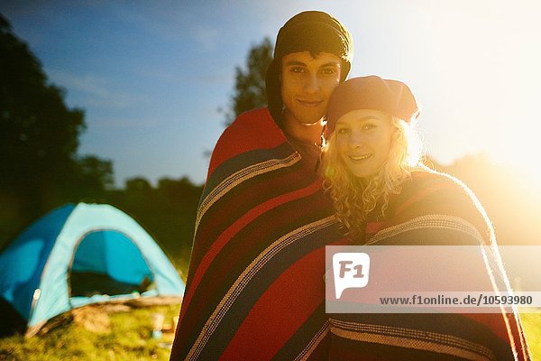 Porträt eines jungen romantischen Campingpaares das bei Sonnenuntergang in eine Decke gehüllt ist.