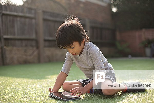 Junge im Garten auf Gras sitzend  mit digitalem Tablett nach unten blickend