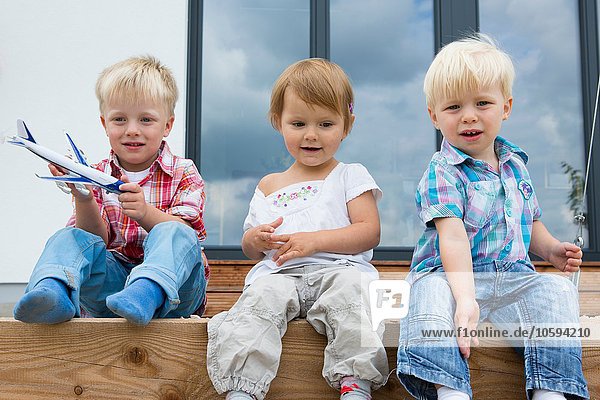 Junge mit Spielzeugflugzeug und zwei Kleinkindern auf der Terrasse sitzend