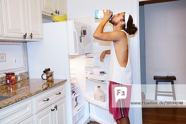 Junger Mann steht neben offenem Kühlschrank und trinkt Milch aus Karton.