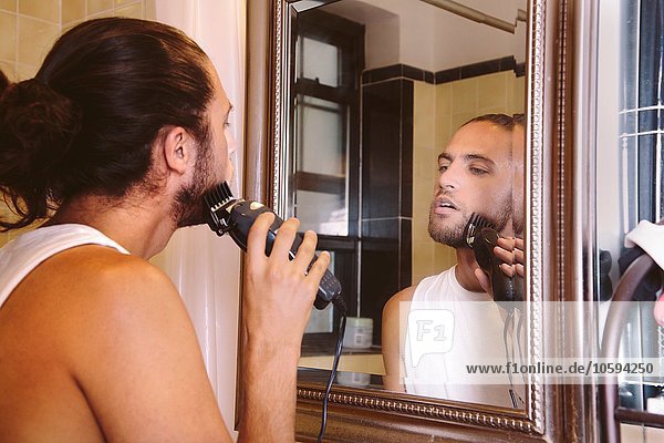 Junger Mann schaut in den Spiegel und rasiert sich mit einem elektrischen Rasierapparat.