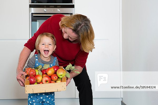 Portrait of female toddler and mother holding crate of apples in kitchen