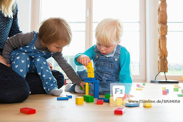 Mittlere erwachsene Frau und zwei Kleinkinder  die mit Bausteinen auf dem Wohnzimmerboden spielen.