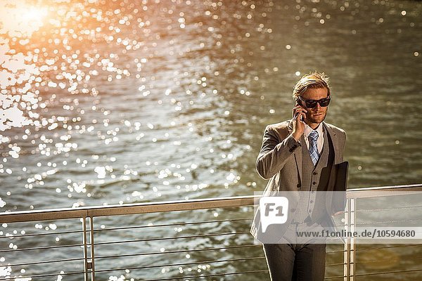 Großer Blickwinkel auf den jungen Geschäftsmann der auf dem Smartphone am Wasser spricht.