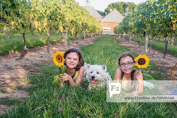 Two young girls lying on grass with pet dog  holding sunflowers