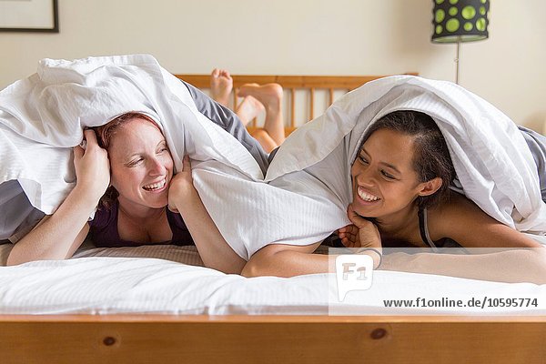 Young women lying on front in bed underneath quilt face to face smiling