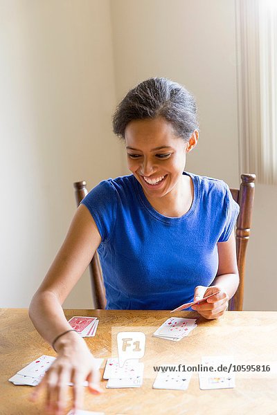 Junge Frau sitzt am Esstisch und spielt lächelnd Karten.