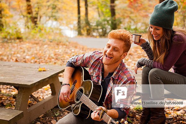 Young couple playing guitar on picnic bench in autumn forest