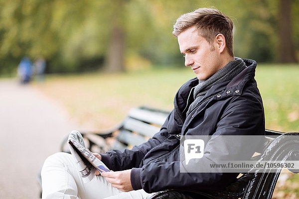 Young man sitting on bench reading newspaper in park