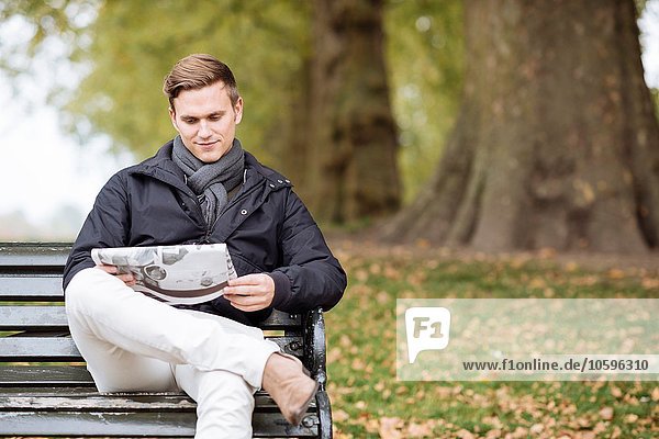 Young man reading newspaper on park bench