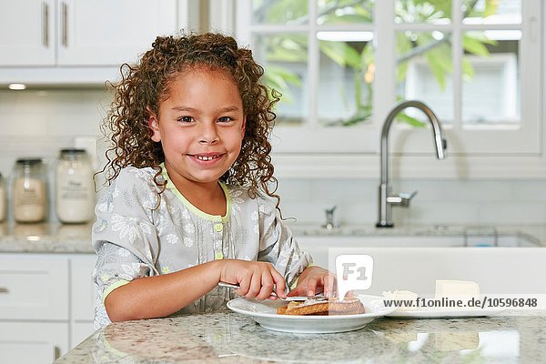 Girl at kitchen counter spreading butter on toast looking at camera smiling