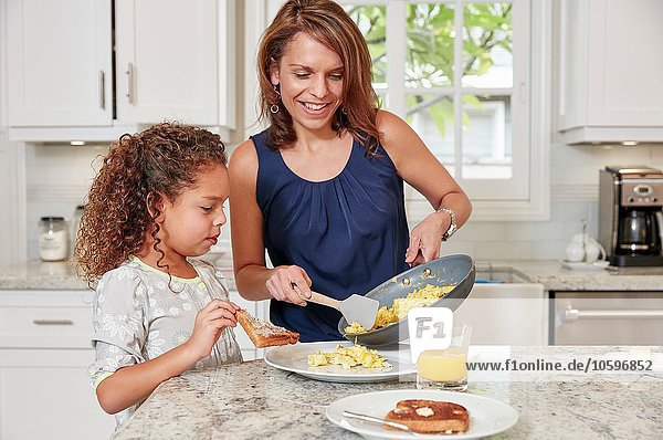 Mother at kitchen counter serving daughter scrambled eggs from frying pan