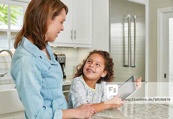 Mother and daughter at kitchen counter using digital tablet  looking up smiling