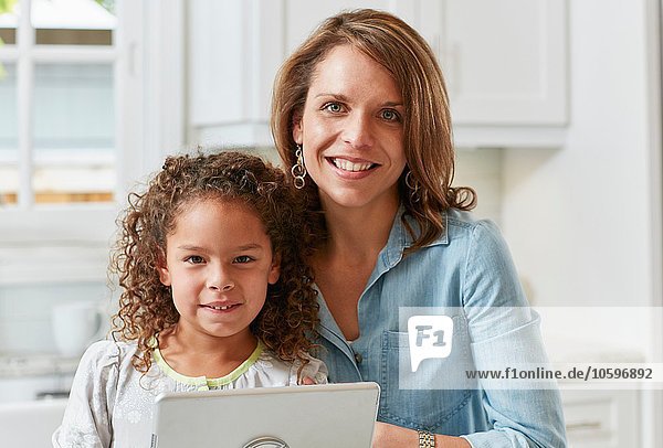 Mutter und Tochter in der Küche mit digitalem Tablett und lächelndem Blick auf die Kamera