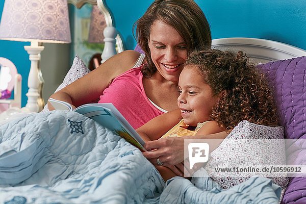 Mother and daughter cuddling in bed reading book smiling