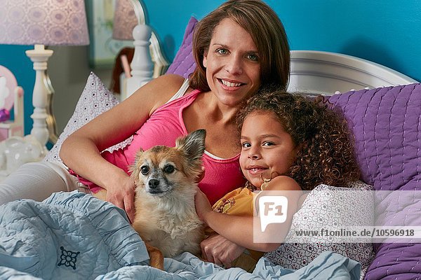 Mother and daughter and dog cuddling in bed looking at camera smiling